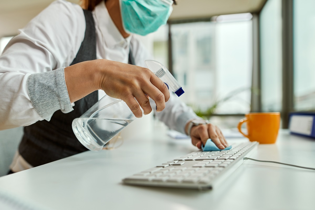closeup de una mujer con tapabocas en oficina limpiando teclado de computador y escritorio